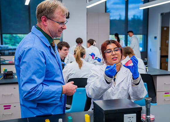 A female student in a lab coat and PPE showing a male instructor how she is using a Petri dish in a lab setting.