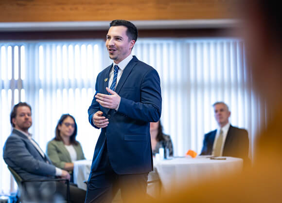A man in suit speaking and gesturing with one hand with several seated people in the background